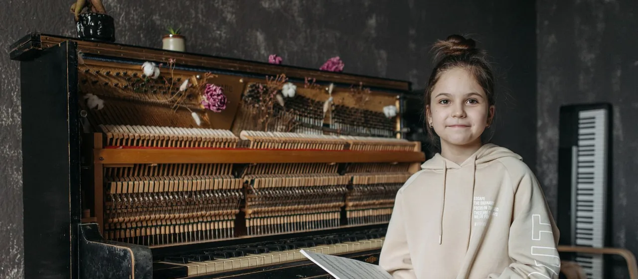 Criança sorrindo com a partitura na mão, ao lado do piano